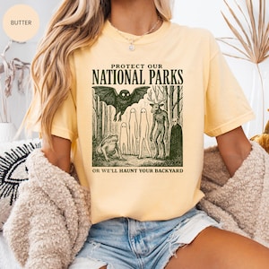 May include: Pale yellow t-shirt featuring the text "PROTECT OUR NATIONAL PARKS" above a forest scene with a bat, ghosts, and a deer-like creature. The text "OR WE'LL HAUNT YOUR BACKYARD" is below the graphic.