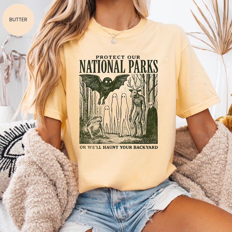 May include: Pale yellow t-shirt featuring the text "PROTECT OUR NATIONAL PARKS" above a forest scene with a bat, ghosts, and a deer-like creature. The text "OR WE'LL HAUNT YOUR BACKYARD" is below the graphic.