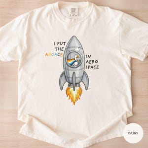 May include: Ivory-colored t-shirt with a graphic of a rocket ship and a duck wearing a cape inside. The text reads "I PUT THE AROACE IN AERO SPACE". The rocket has orange flames coming out of the bottom.