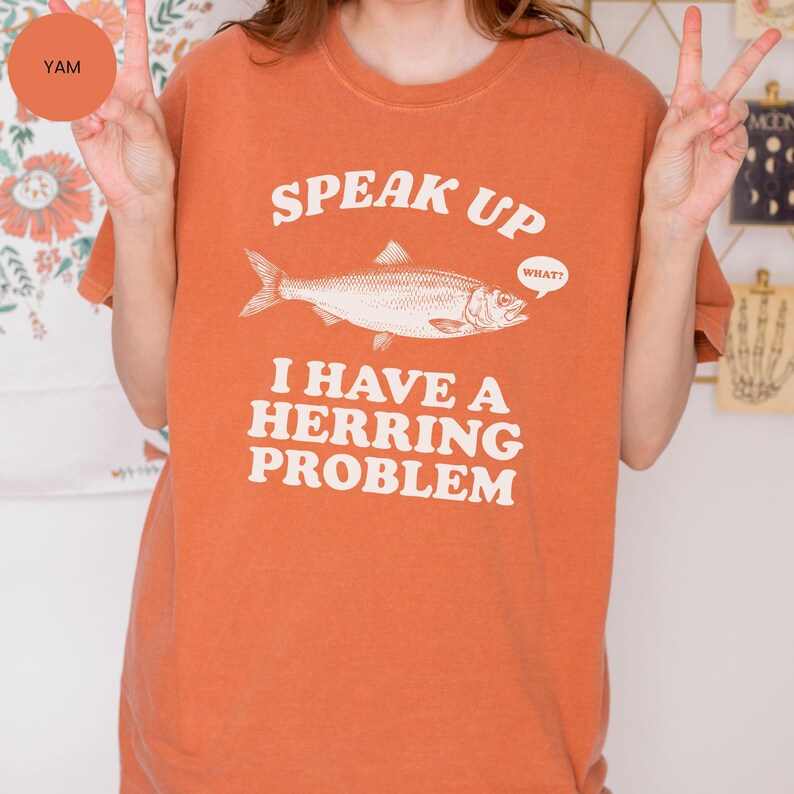 以下が含まれることがあります： オレンジ色のTシャツ。魚のイラストの上に「SPEAK UP」の文字、そして「I HAVE A HERRING PROBLEM」のフレーズがプリントされています。「WHAT？」という吹き出しもあります。Tシャツは単色のオレンジ色です。