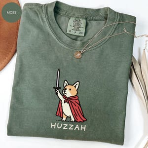 May include: Moss green t-shirt with a graphic of a corgi wearing a red cape and holding a sword. The word "Huzzah" is printed below the corgi. A gold necklace with a pendant is draped on the shirt.