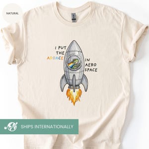May include: Cream-colored t-shirt featuring a cartoon rocket ship with a frog inside, wearing a rainbow flag. The text reads "I PUT THE AROACE IN AERO SPACE". The rocket has orange flames. A fun, whimsical design.