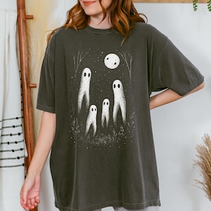 May include: Dark grey t-shirt featuring a whimsical illustration of four white ghost figures under a full moon. The design includes trees and foliage, creating a spooky, yet charming scene. The shirt is a relaxed fit.