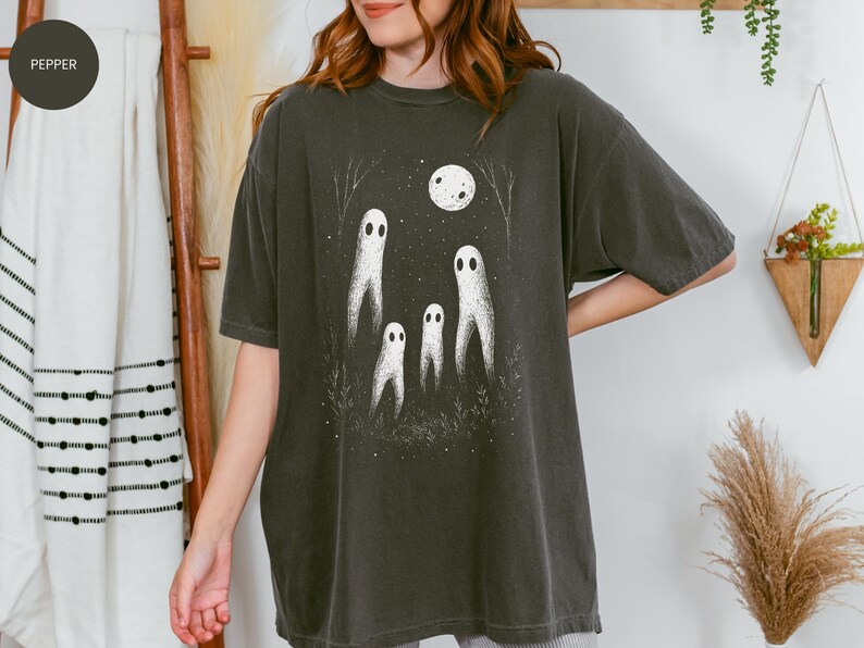 May include: Dark grey t-shirt featuring a whimsical illustration of four white ghost figures under a full moon. The design includes trees and foliage, creating a spooky, yet charming scene. The shirt is a relaxed fit.
