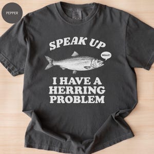 以下が含まれることがあります： 「SPEAK UP I HAVE A HERRING PROBLEM」の文字とニシンの詳細なイラストが描かれた、ペッパーカラーのTシャツ。魚の上にある吹き出しには「WHAT？」と書かれています。