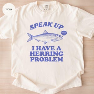 以下が含まれることがあります： アイボリー色のTシャツ。青い文字とグラフィックが特徴です。テキストは「SPEAK UP」と書かれており、その上に青いニシンのイラストがあります。魚の下には「I HAVE A HERRING PROBLEM」と書かれています。吹き出しには「WHAT？」と書かれています。