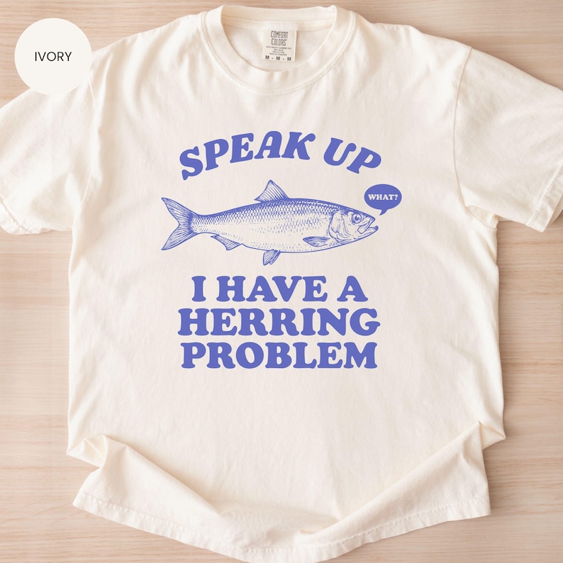 以下が含まれることがあります： アイボリー色のTシャツ。青い文字とグラフィックが特徴です。テキストは「SPEAK UP」と書かれており、その上に青いニシンのイラストがあります。魚の下には「I HAVE A HERRING PROBLEM」と書かれています。吹き出しには「WHAT？」と書かれています。