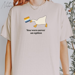 Op de afbeelding: Een wit T-shirt met een cartoon-gans die een regenboogvlag vasthoudt. De tekst "You were never an option" staat onder de gans gedrukt.