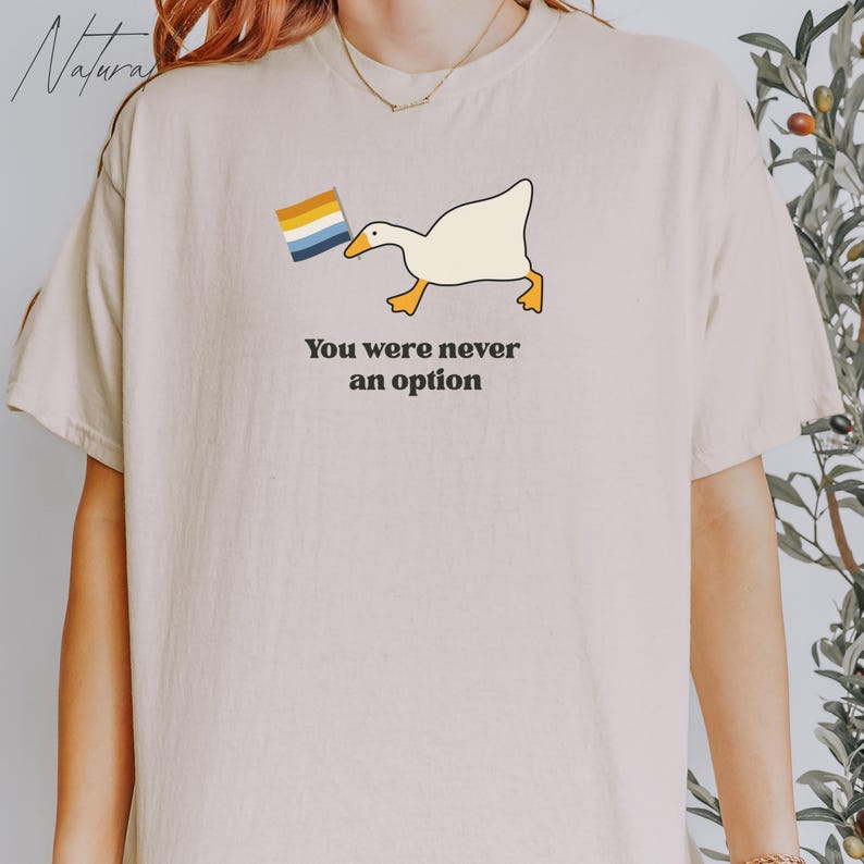 Op de afbeelding: Een wit T-shirt met een cartoon-gans die een regenboogvlag vasthoudt. De tekst "You were never an option" staat onder de gans gedrukt.