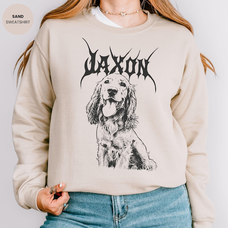 May include: A sand-coloured sweatshirt with a black graphic of a dog and the word "JAXON" above it in a stylized font. The sweatshirt has long sleeves and a crew neck. The text "SAND SWEATSHIRT" is in the upper left corner.