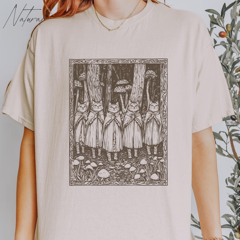 May include: A beige t-shirt with a black and white illustration of a group of cats wearing cloaks and standing in a forest with mushrooms. The illustration is framed with a black border.