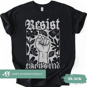 Puede incluir: Una camiseta negra de manga corta con un estampado gráfico blanco de un puño levantado rodeado de flores. El texto "Resist" aparece sobre el puño y el texto "like it's 1968" está impreso debajo en una tipografía estilizada y desgastada.