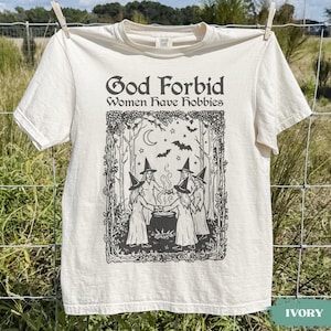 May include: An ivory short-sleeve t-shirt featuring a black graphic print. The text reads "God Forbid Women Have Hobbies" above an illustration of four figures in pointed hats standing in a circle around a steaming cauldron in a forest.