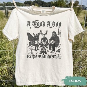 May include: An ivory short-sleeve t-shirt featuring a black graphic print of a moth, a deer-headed figure, and a sasquatch sitting around a campfire reading books. The text reads A BOOK A DAY KEEPS REALITY AWAY in a decorative black gothic font.