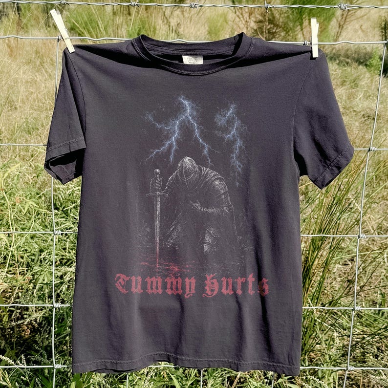 May include: A charcoal gray short-sleeve t-shirt featuring a graphic of a knight in armor kneeling with a sword. Above the knight are blue lightning bolts, and below the graphic is the text "Tummy hurts" printed in a red gothic-style font.