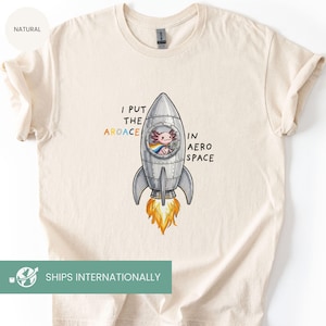 May include: Cream-colored t-shirt featuring a cartoon axolotl in a rocket ship with the text "I put the aroace in aero space." The axolotl is wearing a rainbow scarf. The rocket has flames at the bottom.