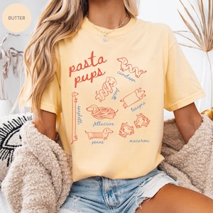 May include: A light yellow t-shirt with the words "pasta pups" in red script. The shirt features illustrations of dachshund dogs representing different pasta shapes, including spaghetti, fettuccine, and lasagna. The word "butter" is in a circle in the upper left corner.