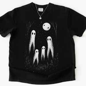 May include: Black t-shirt featuring a whimsical illustration of four white ghost figures under a full moon and starry night sky. The design includes trees and grass, creating a spooky, yet charming scene. A silver necklace with a pendant is visible.
