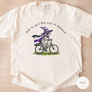 May include: Ivory-colored t-shirt featuring a raccoon riding a bicycle, wearing a purple wizard hat and cape. The text "Off to put the ace in menace" is arched above the raccoon. The shirt is made of a soft material.
