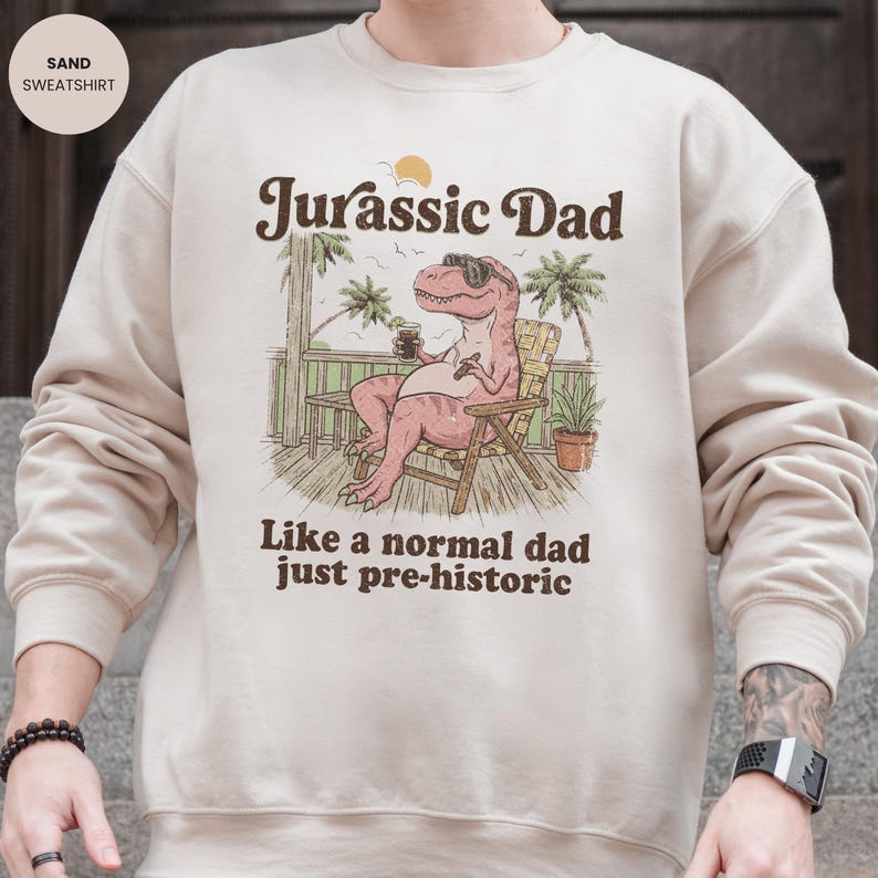 May include: Sand-colored sweatshirt with the text "Jurassic Dad" and an illustration of a dinosaur wearing sunglasses and relaxing in a chair. The text "Like a normal dad just pre-historic" is below the illustration.