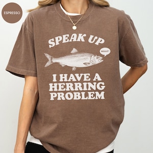 以下が含まれることがあります： エスプレッソ色のTシャツ。魚のイラストの上に「SPEAK UP」、そして「I HAVE A HERRING PROBLEM」というフレーズがプリントされています。「WHAT？」という吹き出しがあります。ゆったりとしたフィット感のシャツです。