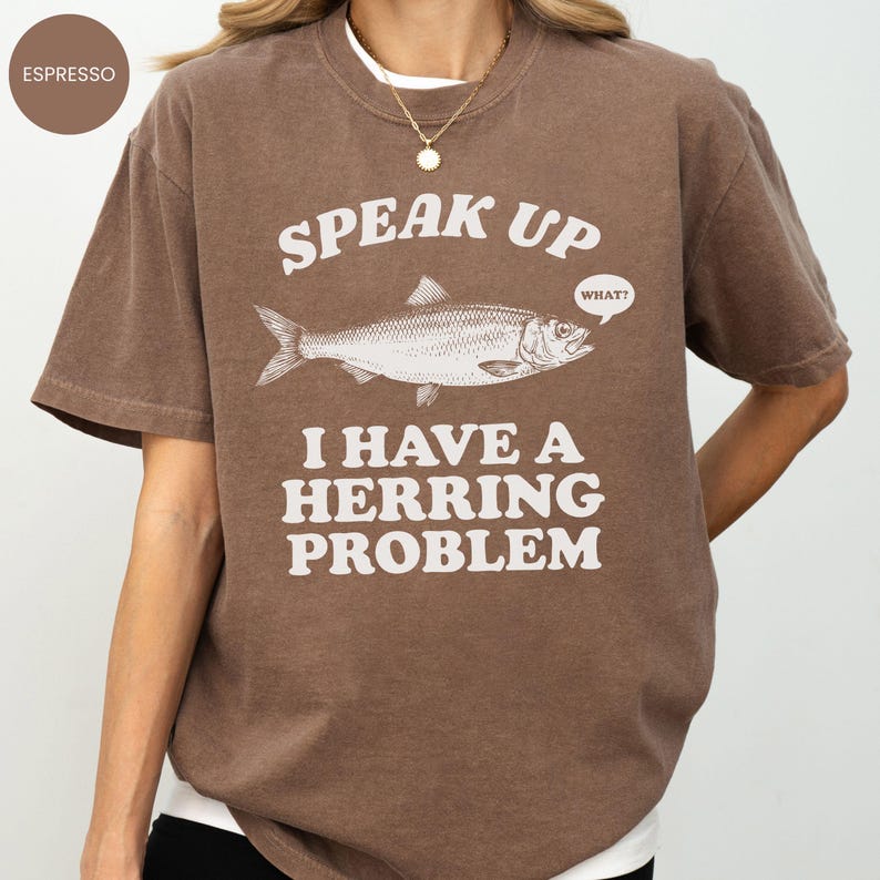以下が含まれることがあります： エスプレッソ色のTシャツ。魚のイラストの上に「SPEAK UP」、そして「I HAVE A HERRING PROBLEM」というフレーズがプリントされています。「WHAT？」という吹き出しがあります。ゆったりとしたフィット感のシャツです。