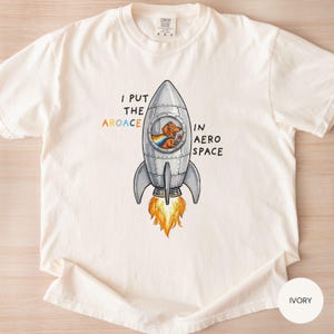 May include: Ivory-colored t-shirt featuring a cartoon rocket ship with a dachshund pilot wearing a rainbow cape. The text reads "I put the aroace in aerospace". The rocket has orange flames and the shirt has a crew neck.