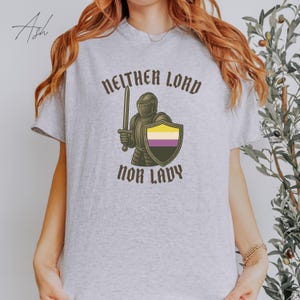 May include: A light gray t-shirt featuring the text "NEITHER LORD NOR LADY" in a gothic font. The graphic includes a knight holding a sword and a shield with the non-binary pride flag colors. The shirt is a casual wear item.