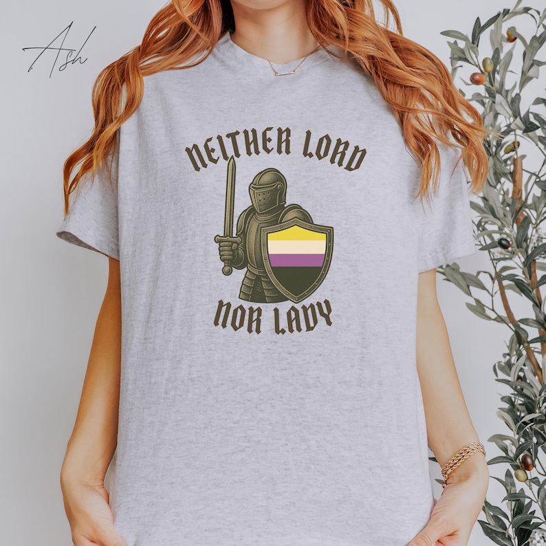 May include: A light gray t-shirt featuring the text "NEITHER LORD NOR LADY" in a gothic font. The graphic includes a knight holding a sword and a shield with the non-binary pride flag colors. The shirt is a casual wear item.
