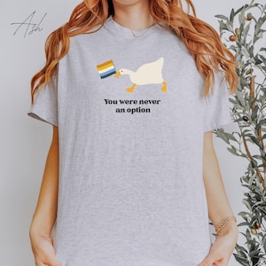 Op de afbeelding: Een grijs T-shirt met een witte gans die een regenboogvlag in zijn bek houdt. De tekst "You were never an option" staat onder de gans.