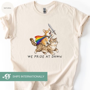 May include: Cream-colored t-shirt featuring a whimsical illustration of a corgi wearing a rainbow cape and holding a sword, riding on a tabby cat. The text "WE PRIDE AT DAWN" is printed below the image.