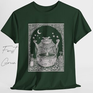 May include: A forest green t-shirt with a white graphic of a frog in a robe stirring a cauldron under a crescent moon and stars. The graphic is surrounded by a decorative frame.