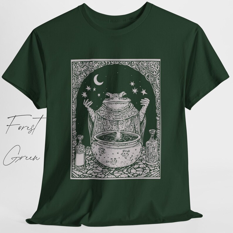 May include: A forest green t-shirt with a white graphic of a frog in a robe stirring a cauldron under a crescent moon and stars. The graphic is surrounded by a decorative frame.