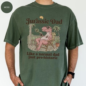 May include: Moss green t-shirt with the text "Jurassic Dad" above an illustration of a relaxed dinosaur wearing sunglasses and holding a drink. The text "Like a normal dad just pre-historic" is below the illustration.