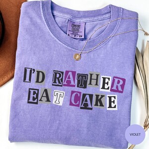 May include: A violet-colored t-shirt with the phrase "I'd Rather Eat Cake" in a block letter design. A gold necklace with a pendant rests on the shirt. The shirt has a Comfort Colors tag.