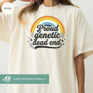 May include: Cream-colored t-shirt with a rainbow graphic and the text "Proud genetic dead end" in a black, blue, and white design. The shirt has short sleeves and a relaxed fit.