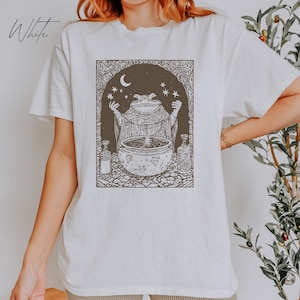 May include: White t-shirt with a graphic print of a frog in a robe holding a cauldron with stars and a crescent moon in the background.