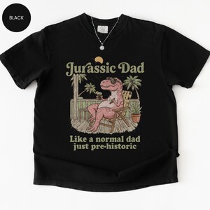May include: Black t-shirt with a graphic of a pink Tyrannosaurus Rex wearing sunglasses, sitting in a chair, and holding a drink. Text reads "Jurassic Dad" above and "Like a normal dad just pre-historic" below.