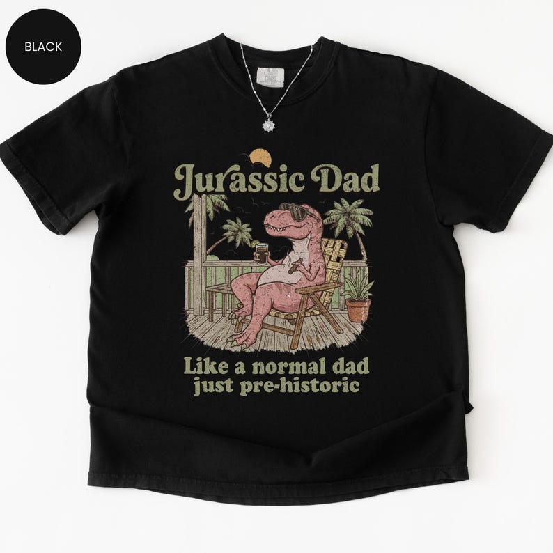 May include: Black t-shirt with a graphic of a pink Tyrannosaurus Rex wearing sunglasses, sitting in a chair, and holding a drink. Text reads "Jurassic Dad" above and "Like a normal dad just pre-historic" below.