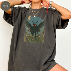 May include: Dark grey t-shirt featuring a whimsical illustration of a moth-like creature with red eyes, set against a night sky with a crescent moon and flowers. The graphic is centered on the front of the shirt.