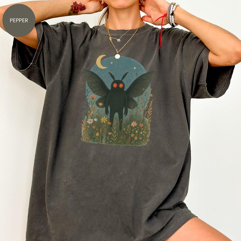 May include: Dark grey t-shirt featuring a whimsical illustration of a moth-like creature with red eyes, set against a night sky with a crescent moon and flowers. The graphic is centered on the front of the shirt.