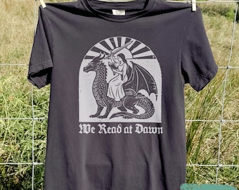 Puede incluir: Una camiseta gris carbón de manga corta con un estampado gráfico gris claro de una persona sentada sobre un dragón mientras sostiene una espada y un libro. El diseño está dentro de un arco semicircular con rayos de sol y el texto We Read at Dawn.