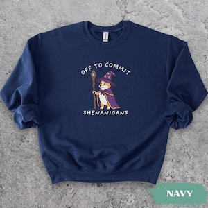 Könnte beinhalten: Ein marineblaues Sweatshirt mit Rundhalsausschnitt und einer Grafik eines Corgis, der als Zauberer mit violettem Hut und Umhang verkleidet ist und einen Holzstab hält. Der Text "OFF TO COMMIT SHENANIGANS" steht in Weiß über und unter dem Motiv.
