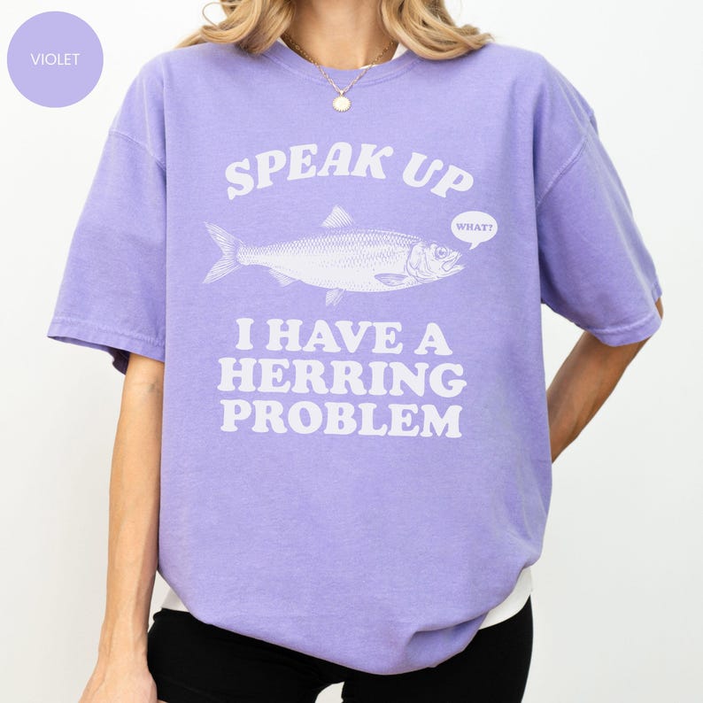 以下が含まれることがあります： 紫色のTシャツ。魚のイラストの上に「SPEAK UP」の文字、そして「I HAVE A HERRING PROBLEM」というフレーズがプリントされています。「WHAT？」という吹き出しもあります。半袖のシャツです。