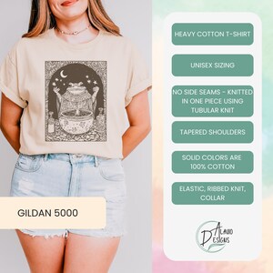 May include: A beige t-shirt with a graphic of a frog holding a cauldron with stars and a crescent moon. The text "GILDAN 5000" is on the bottom of the shirt. The shirt is made of heavy cotton and has a unisex sizing. It is knitted in one piece using tubular knit and has tapered shoulders. The shirt is 100% cotton and has an elastic, ribbed knit collar.