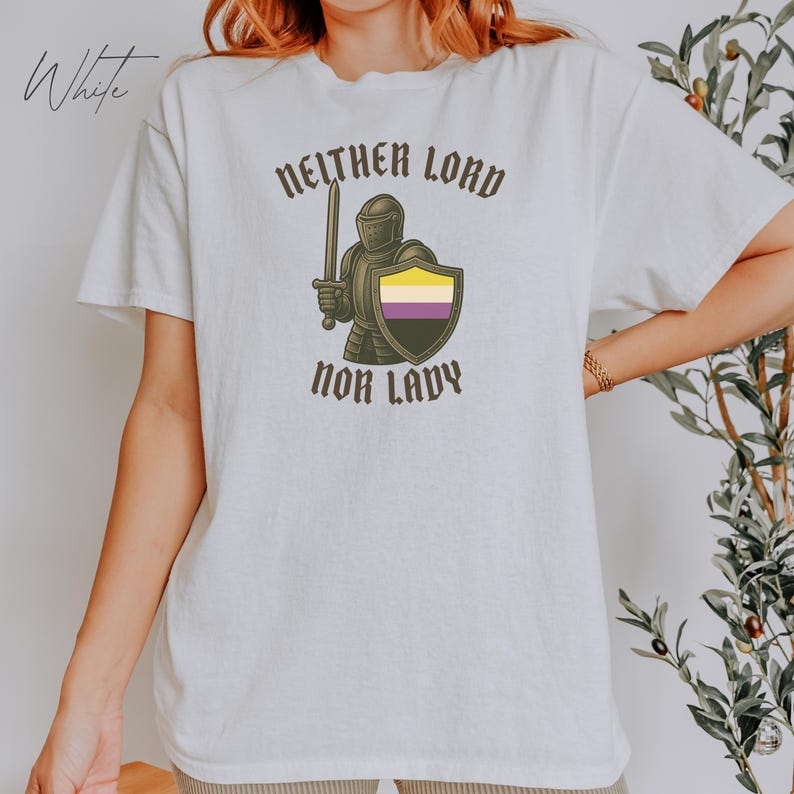 May include: White t-shirt with the text "Neither Lord Nor Lady" and a graphic of a knight holding a sword and shield. The shield features the non-binary pride flag colors: yellow, white, purple, and black. The word "White" is written in cursive in the upper left corner.
