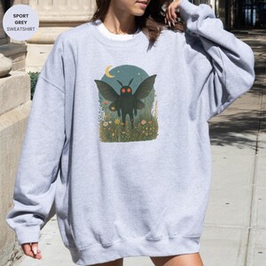 May include: Sport grey sweatshirt featuring a whimsical illustration of a moth-like creature with large wings, standing in a field of flowers under a crescent moon. The design is set against a teal background. The sweatshirt is a comfortable, casual style.