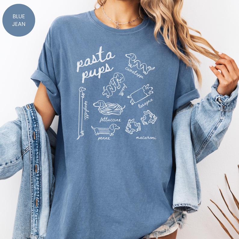 May include: Blue jean colored t-shirt with the words "pasta pups" in white script. The shirt features white line drawings of dachshunds representing different pasta shapes, including spaghetti, cavatappi, and lasagna.