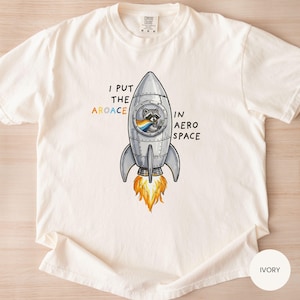 May include: Ivory-colored t-shirt featuring a rocket ship with a raccoon pilot and the text "I put the aroace in aerospace." The rocket has orange flames and a rainbow flag. The shirt is a Comfort Colors brand.