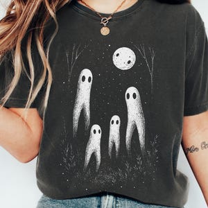 May include: Graphite-colored t-shirt featuring a whimsical illustration of four white ghost figures under a full moon and starry night sky. The design includes thin tree silhouettes and grass, creating a spooky, yet charming scene.